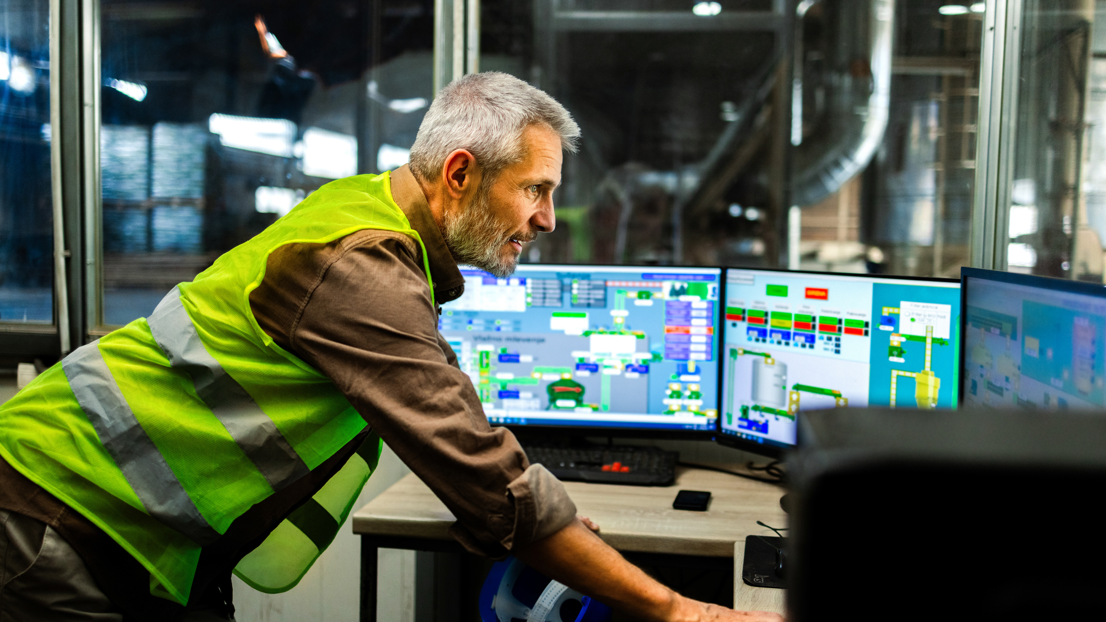 A worker is leaning forward on a desk and examining multiple computer monitors displaying various data and control panels in an industrial office.