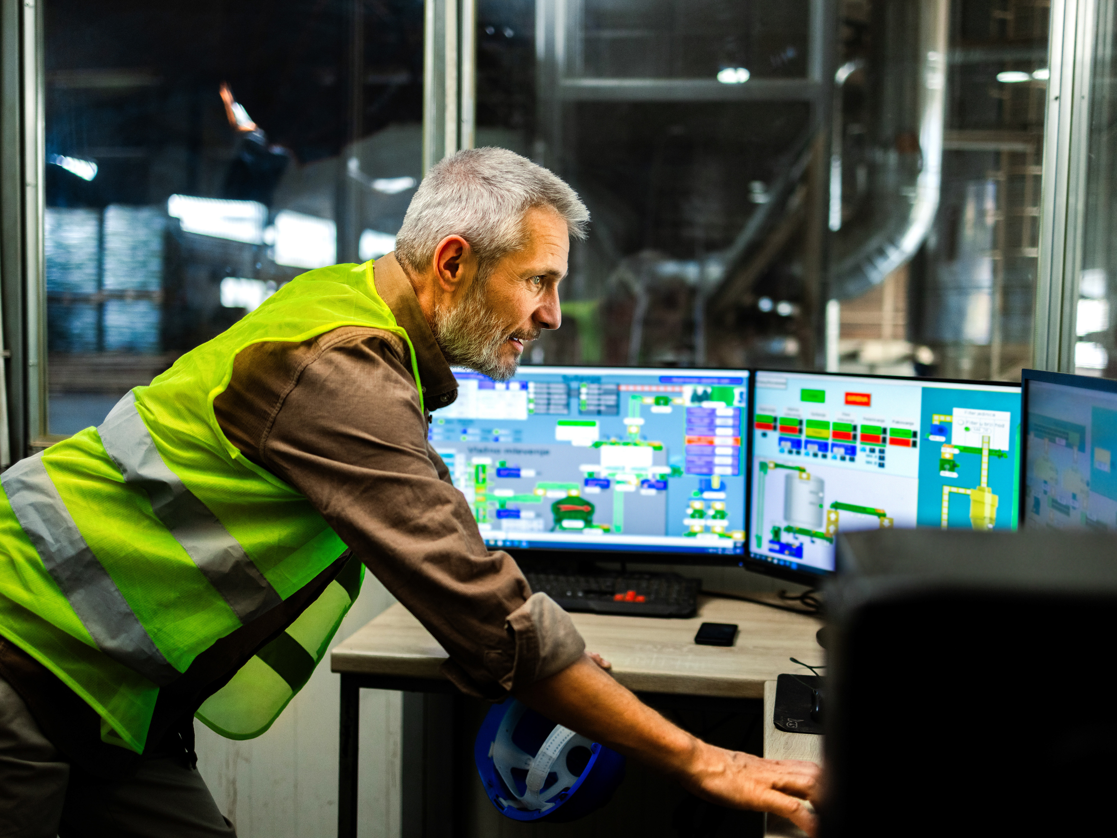 A worker is leaning forward on a desk and examining multiple computer monitors displaying various data and control panels in an industrial office.