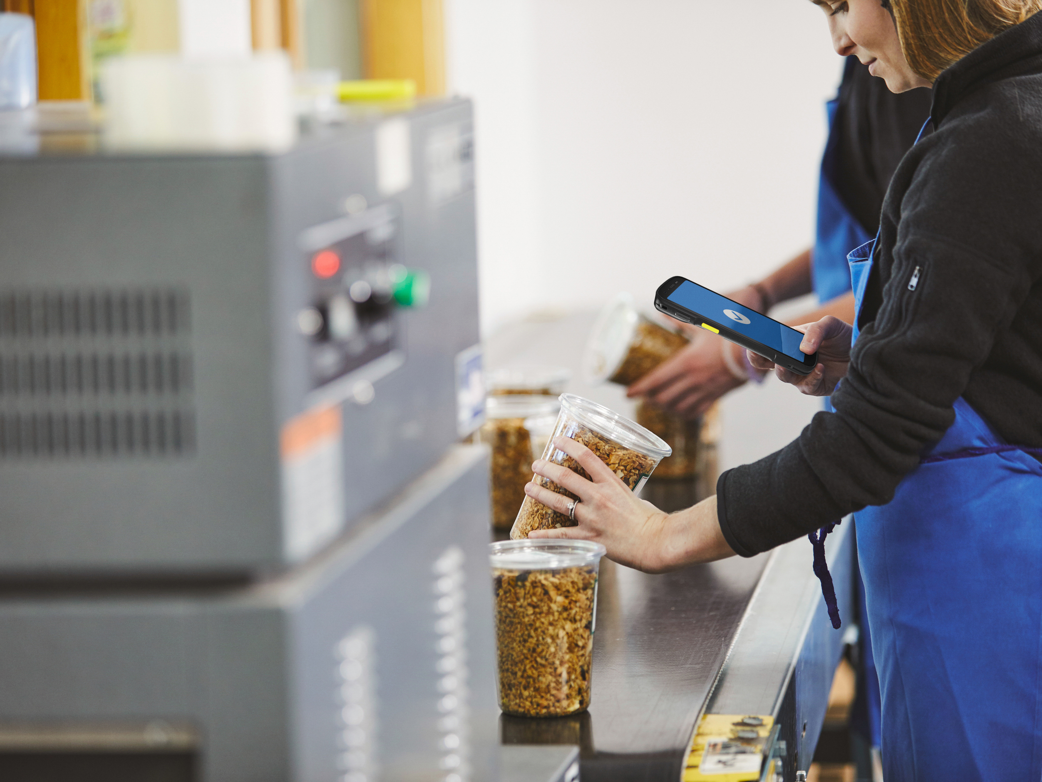 A worker using a handheld mobile computer to scan food products on a conveyor belt inside of a manufacturing plant.