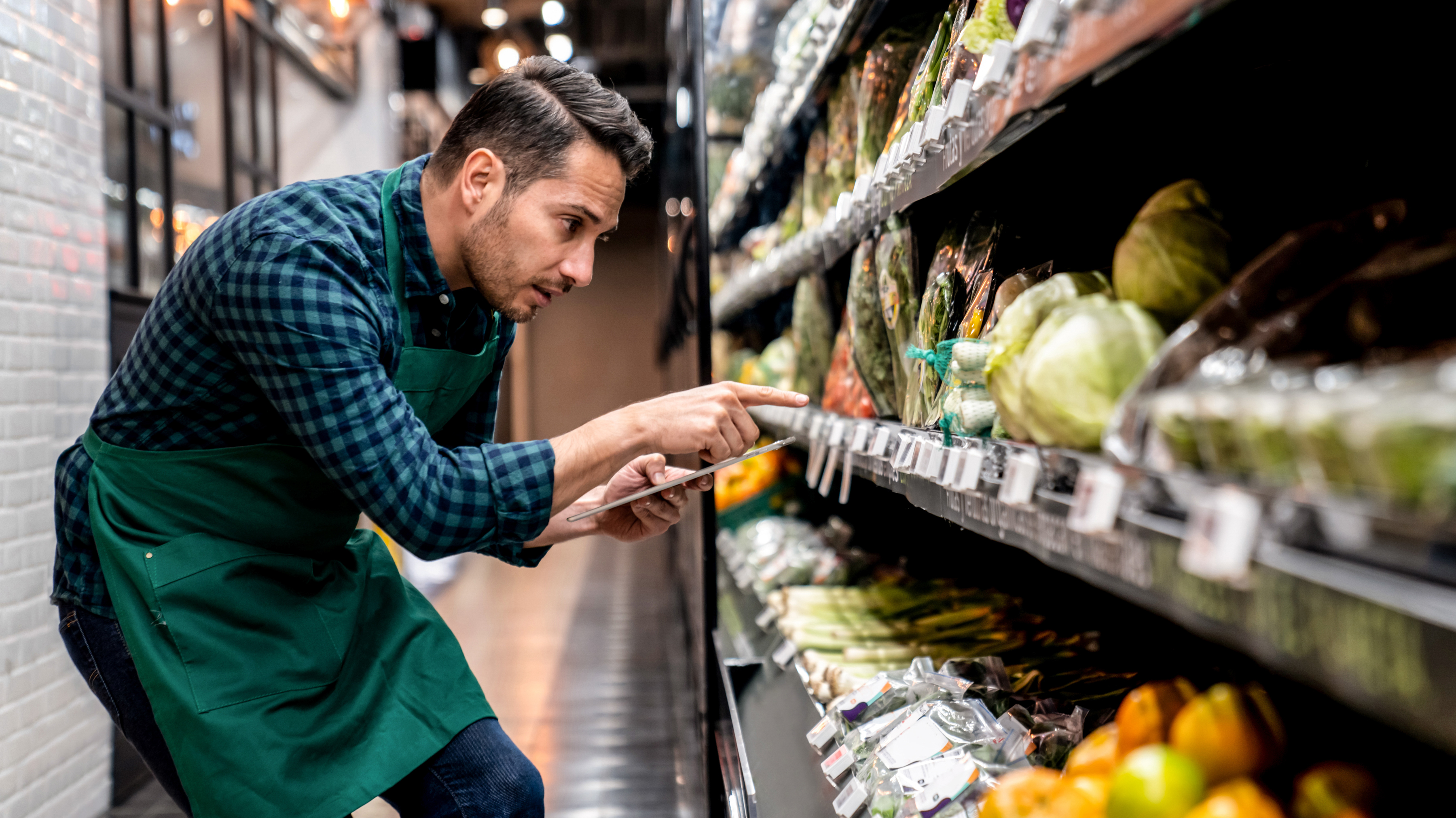 A worker  is crouching and pointing at a shelf label as he checks the produce inventory in a grocery store.