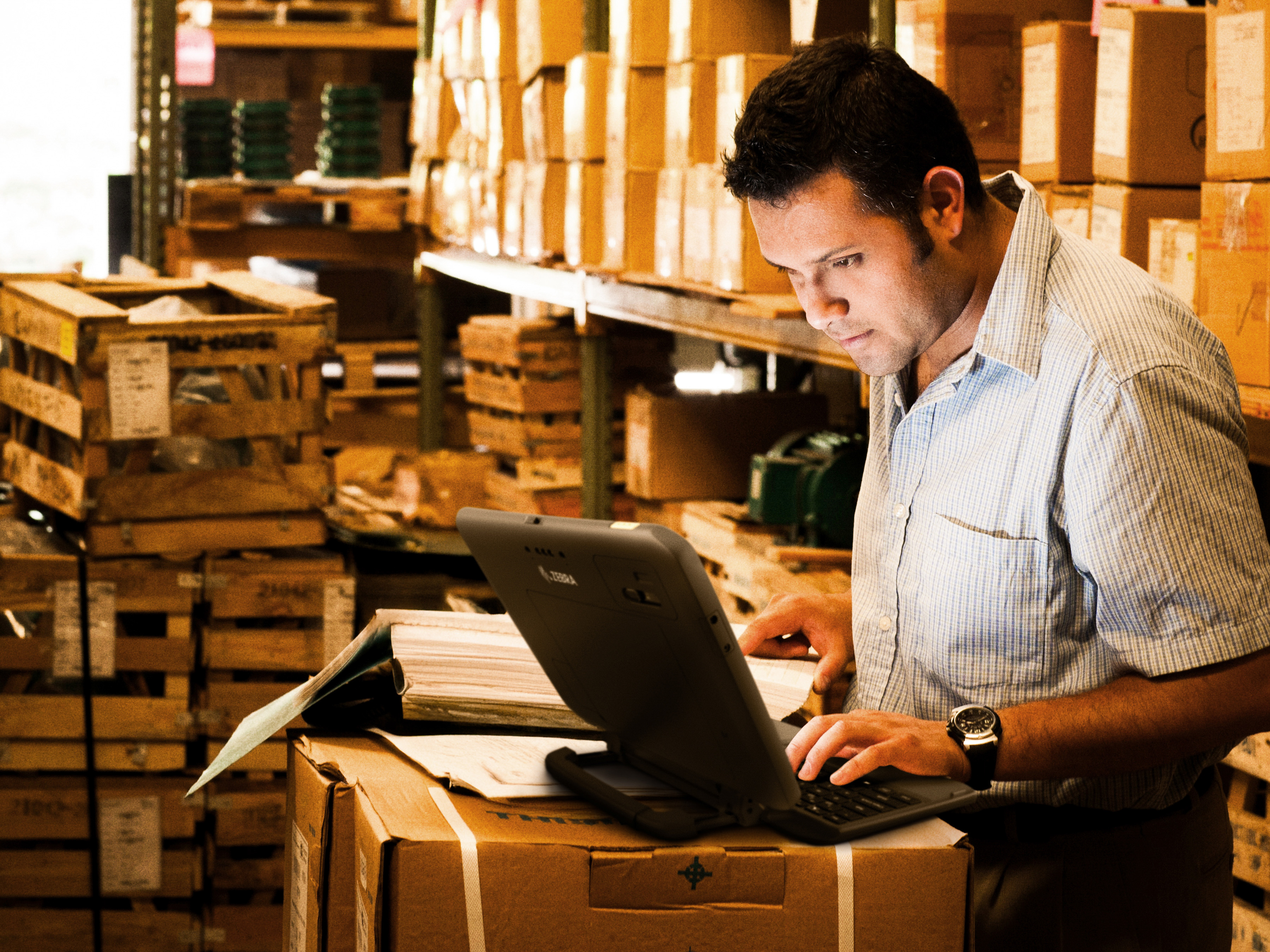 A back-of-house employee is working on a laptop while reviewing inventory stock in a store room filled with shelves of cardboard boxes and wooden crates.