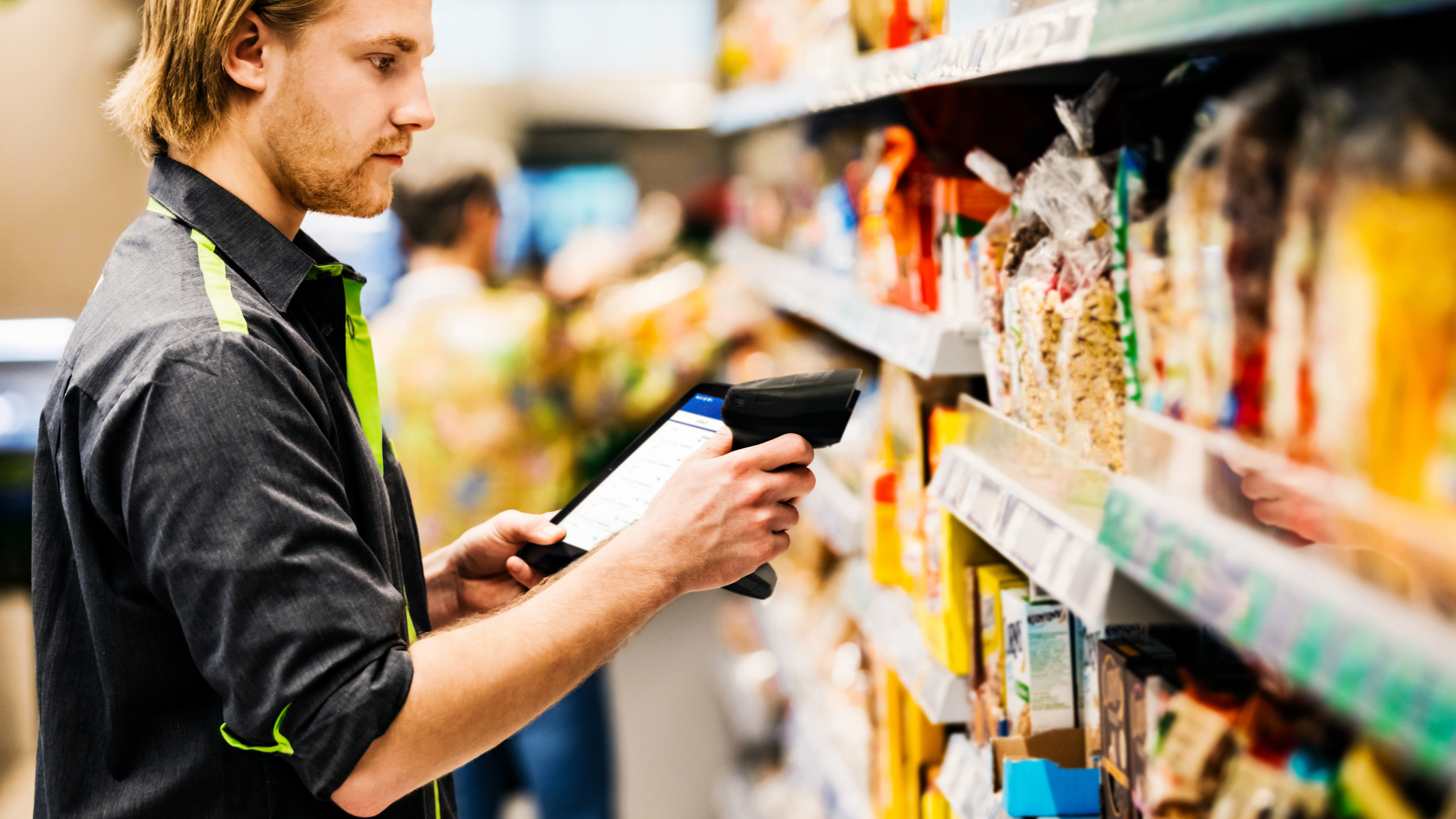 A front-of-house retail employee using a handheld barcode scanner and mobile computer to scan stock on a grocery store shelf.