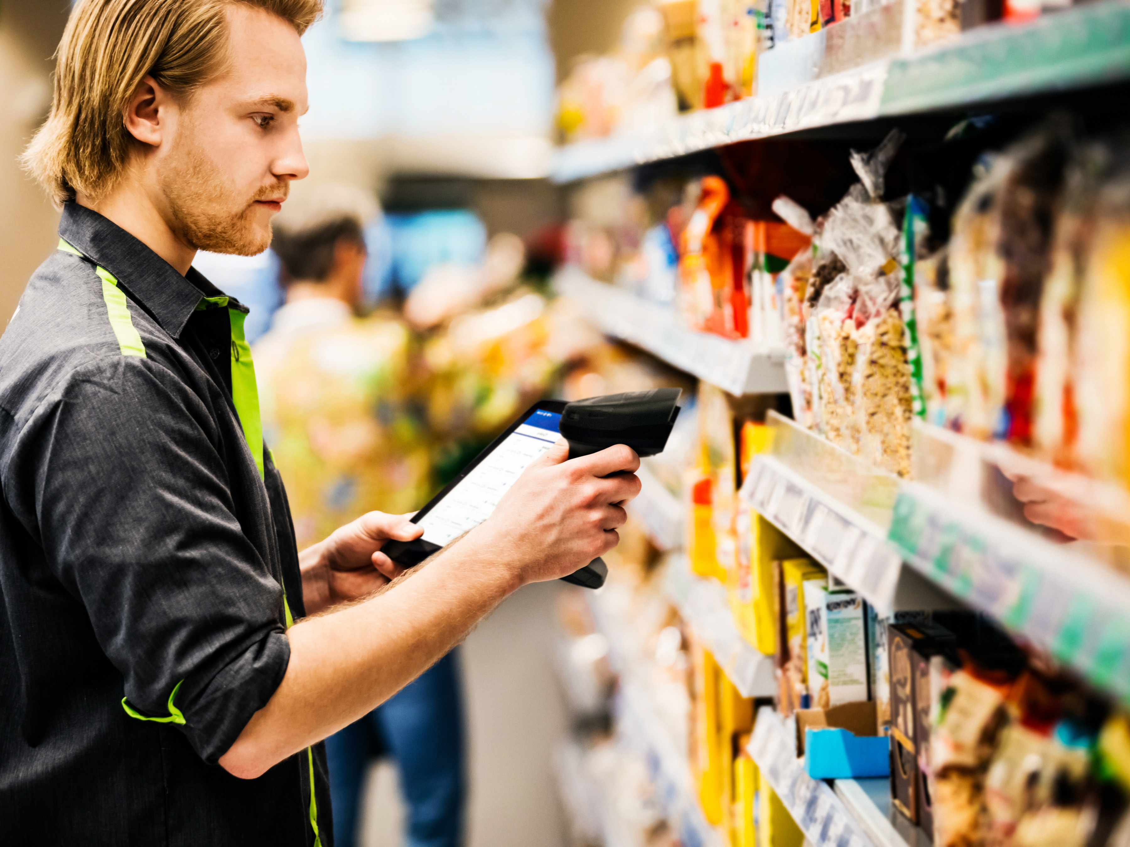 A front-of-house retail employee using a handheld barcode scanner and mobile computer to scan stock on a grocery store shelf.