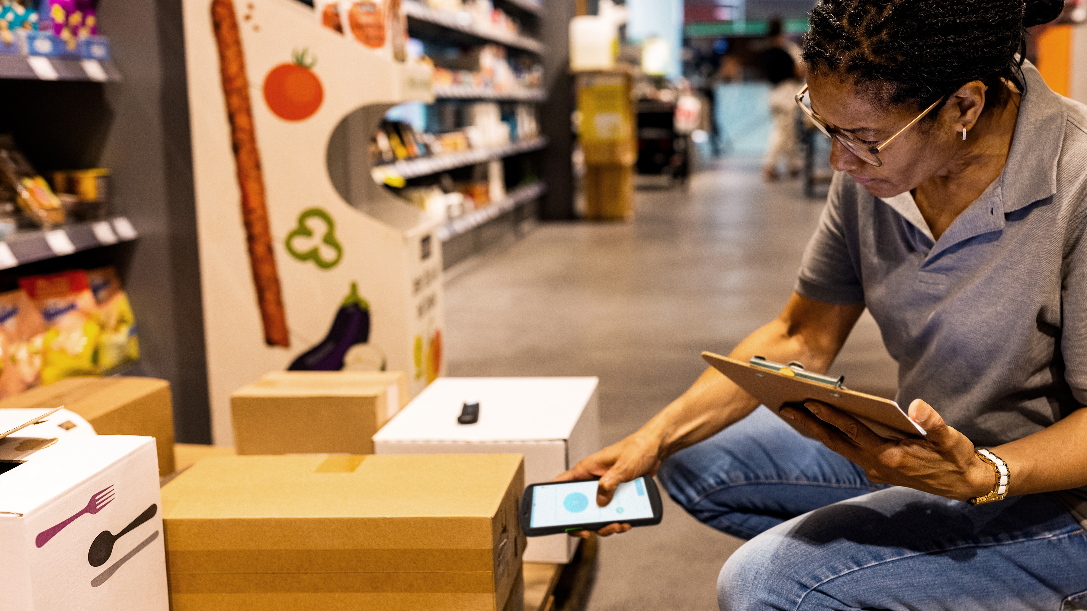 An employee holding a tablet scans stock boxes with a handheld scanner inside a grocery store aisle.