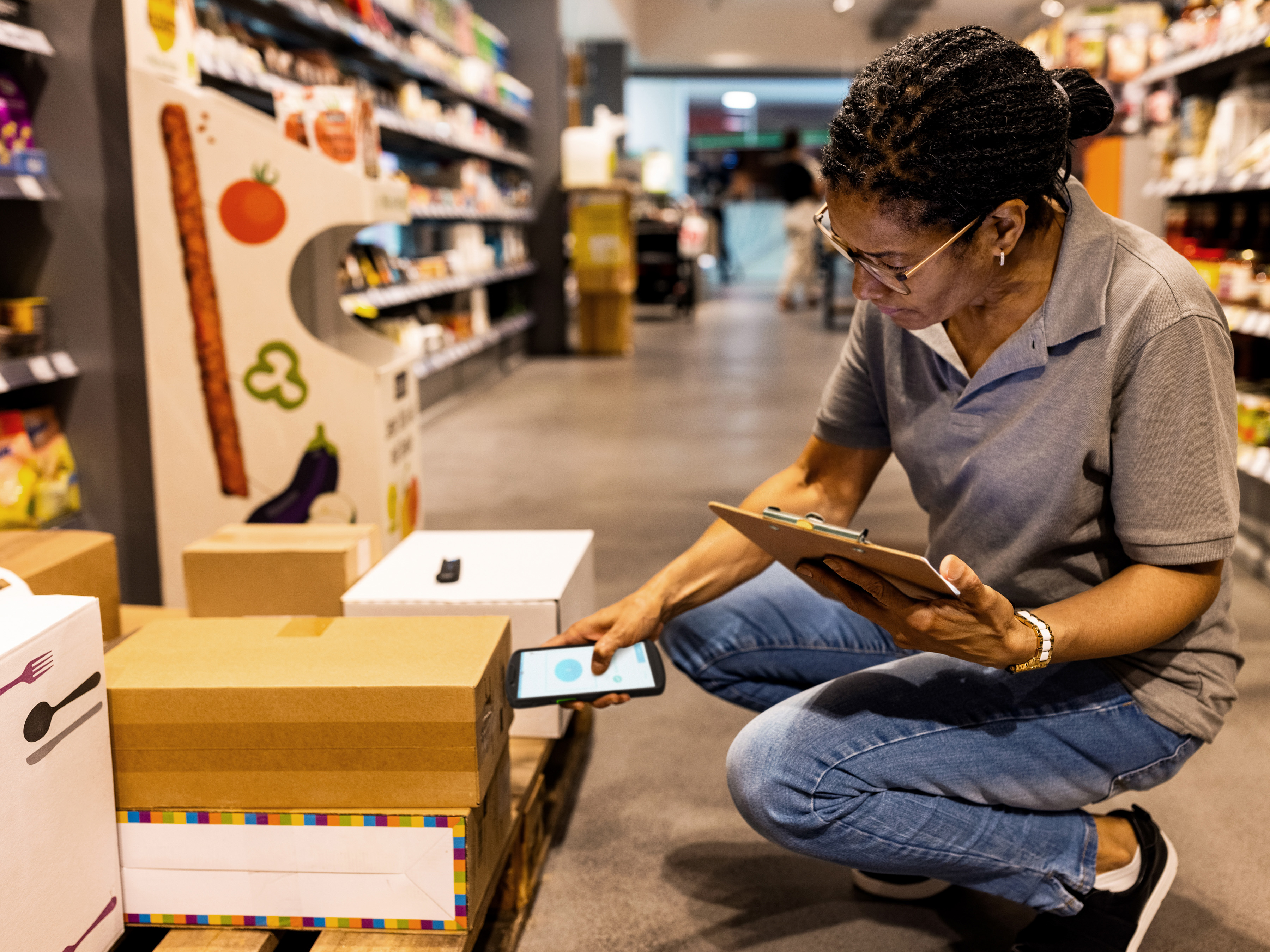An employee holding a tablet scans stock boxes with a handheld scanner inside a grocery store aisle.