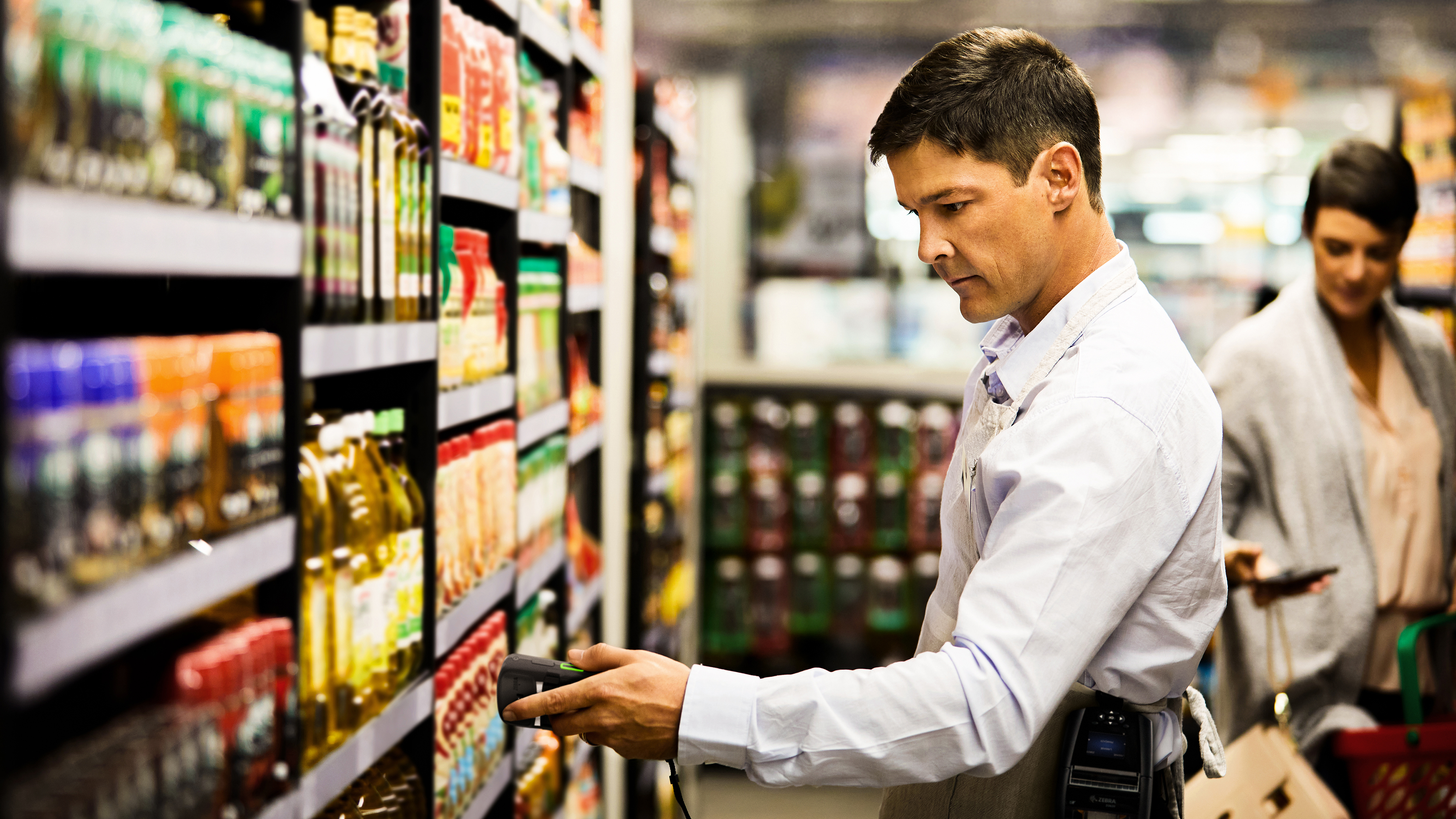 Employee scanning grocery products on shelf aisle using a tablet and handheld scanner.