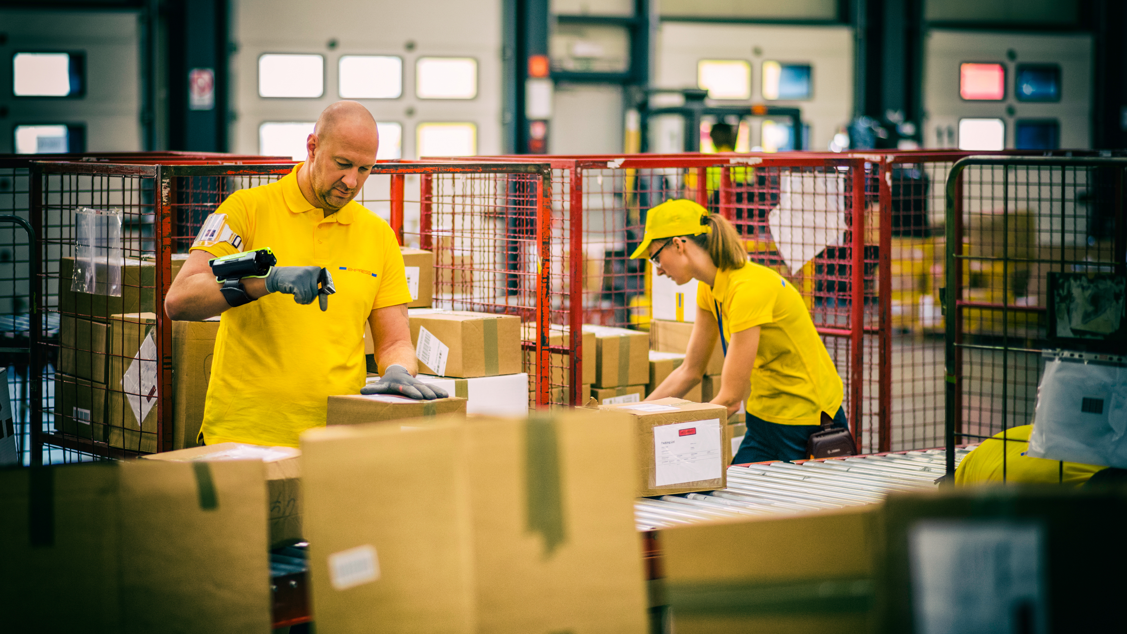 Two postal workers in yellow uniforms are sorting and scanning boxes using wearable scanners.