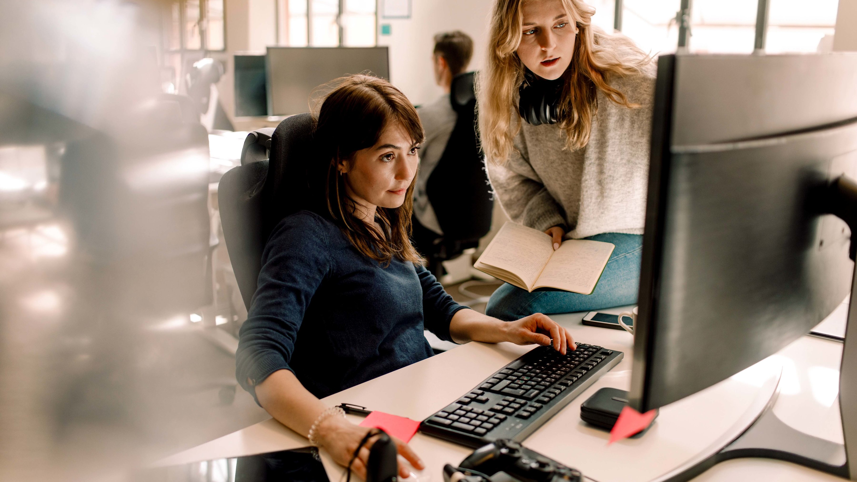 Two colleagues are discussing a business project and working together at a desktop computer in the office, with one sitting and typing while the other stands holding an open notebook.