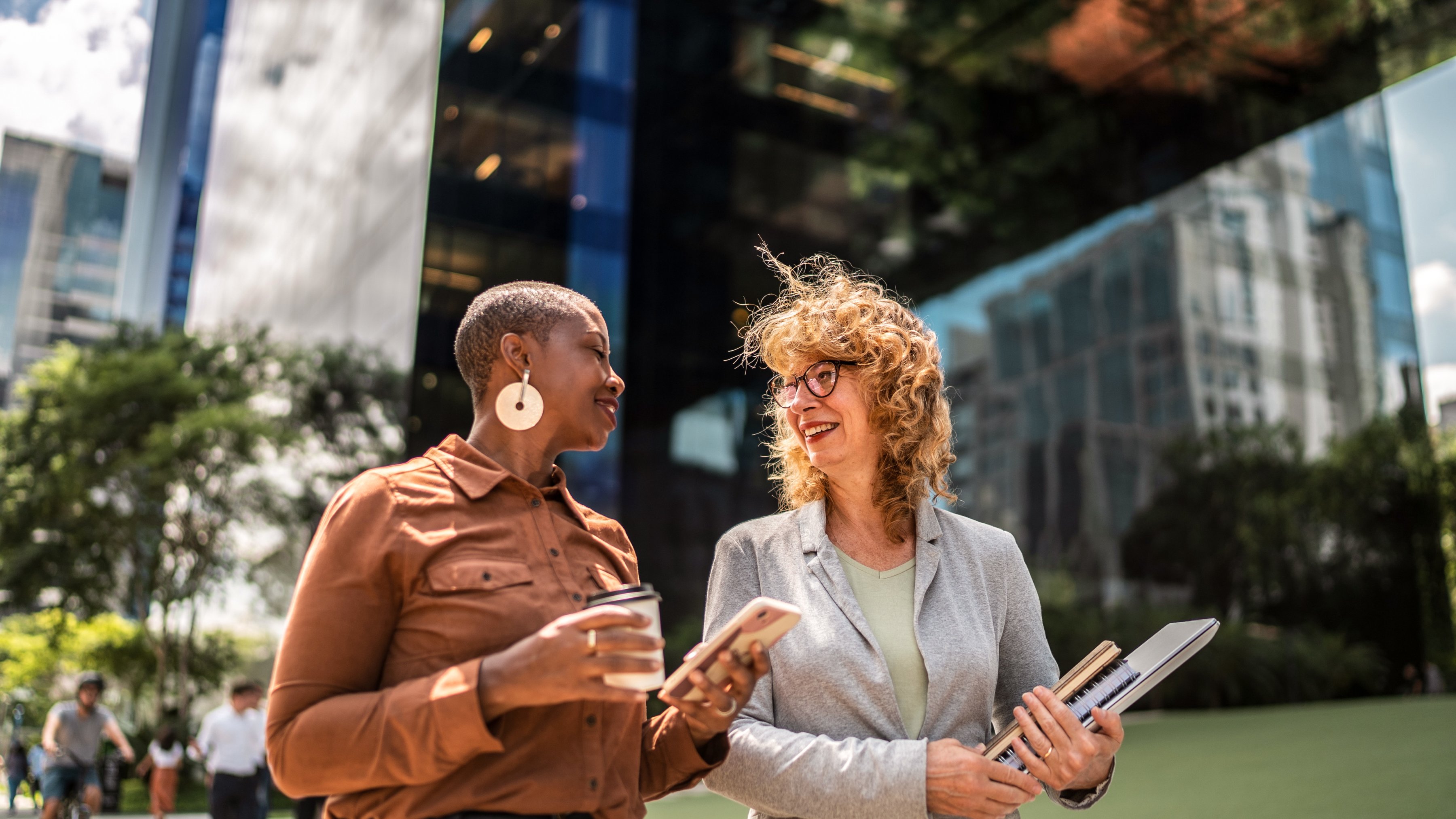 Two colleagues are walking outside and having a conversation, while one holds a mobile computer and the other holds a laptop and documents.