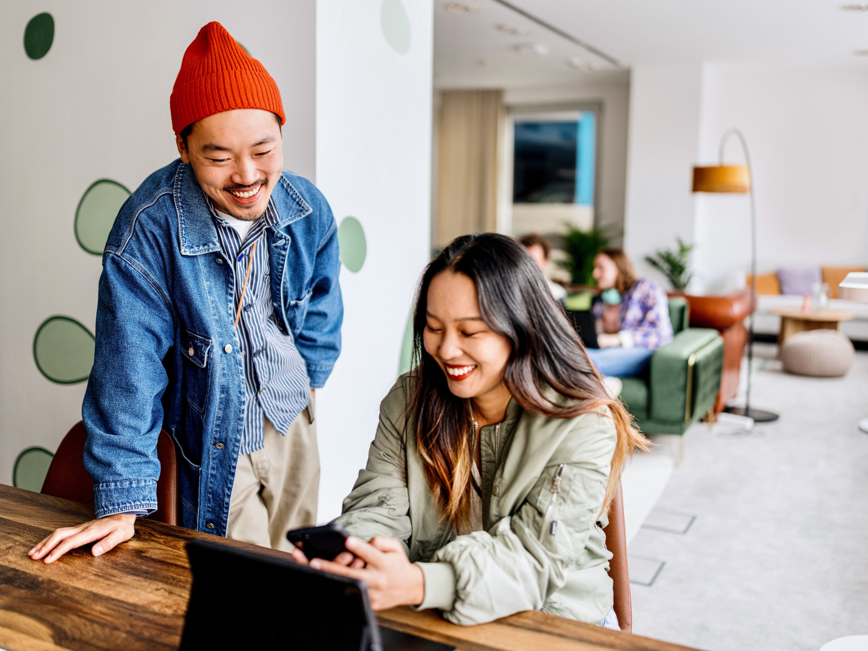 Two colleagues are engaged in a discussion at a table, with one person standing and leaning on the table while the other sits and shows the screen display on a mobile computer. 