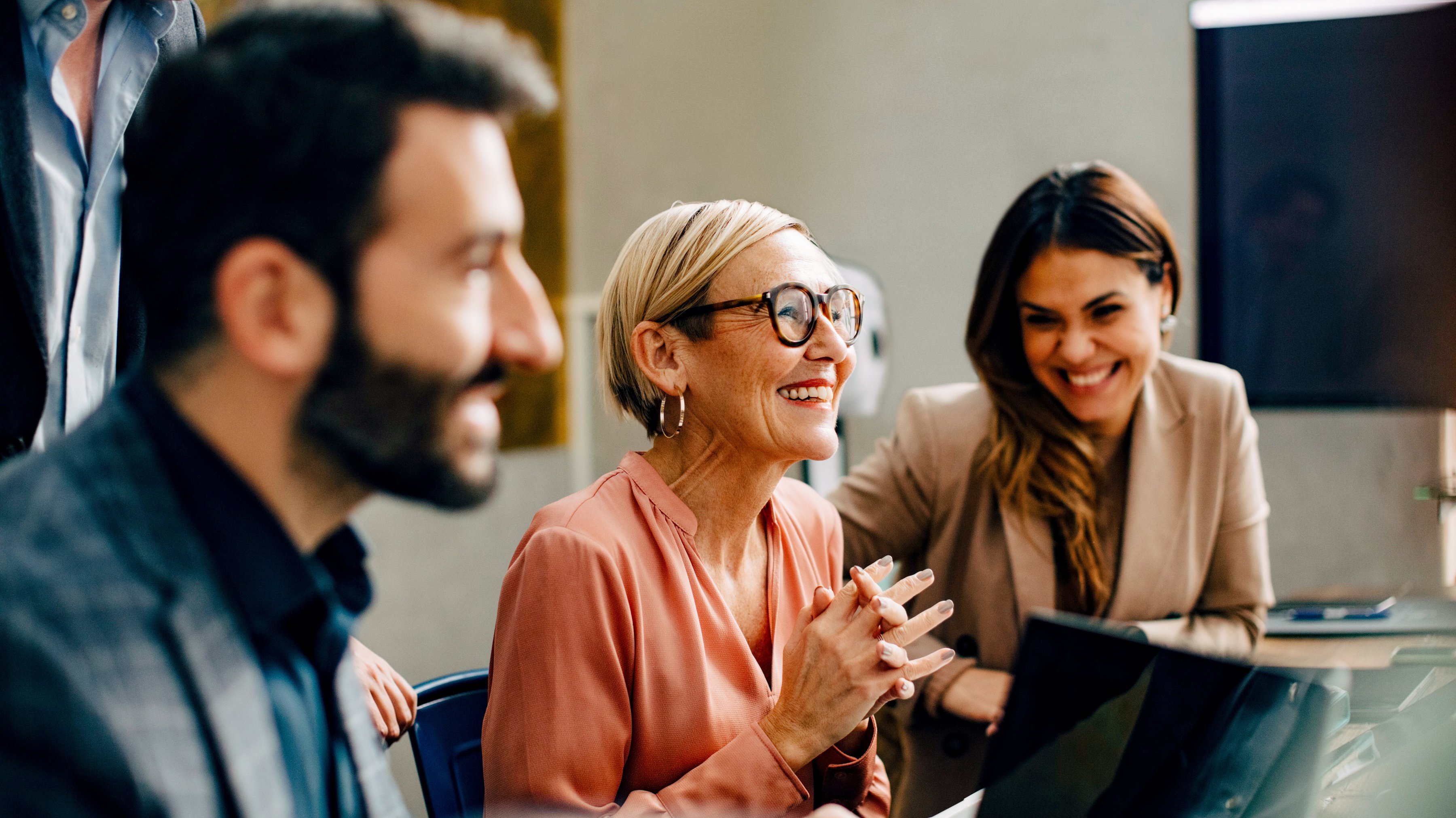 A group of colleagues in business attire are gathered around a computer, reacting positively to a speaker during a meeting in the office.