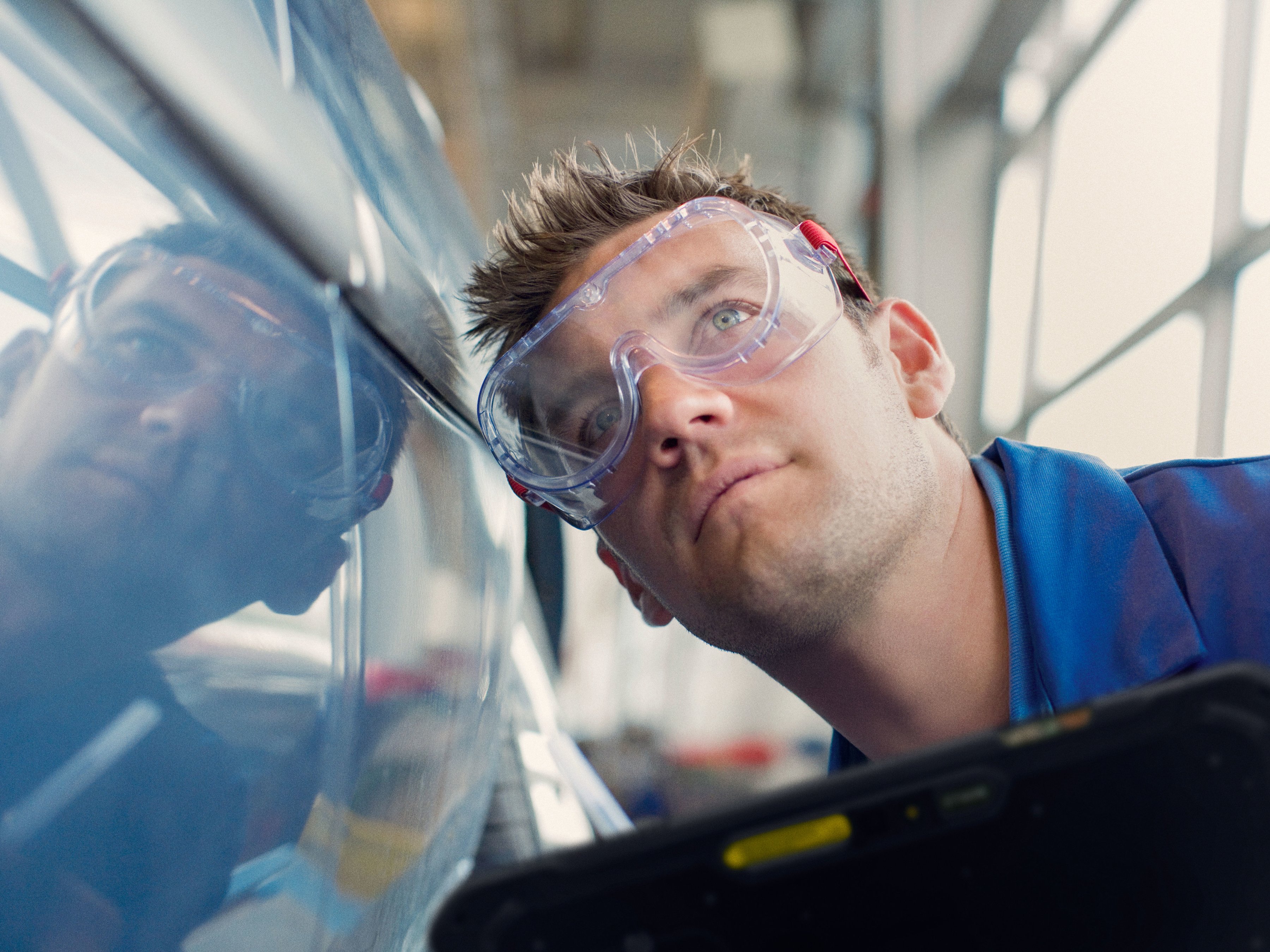 A technician wearing safety goggles is closely inspecting a car at a production plant.