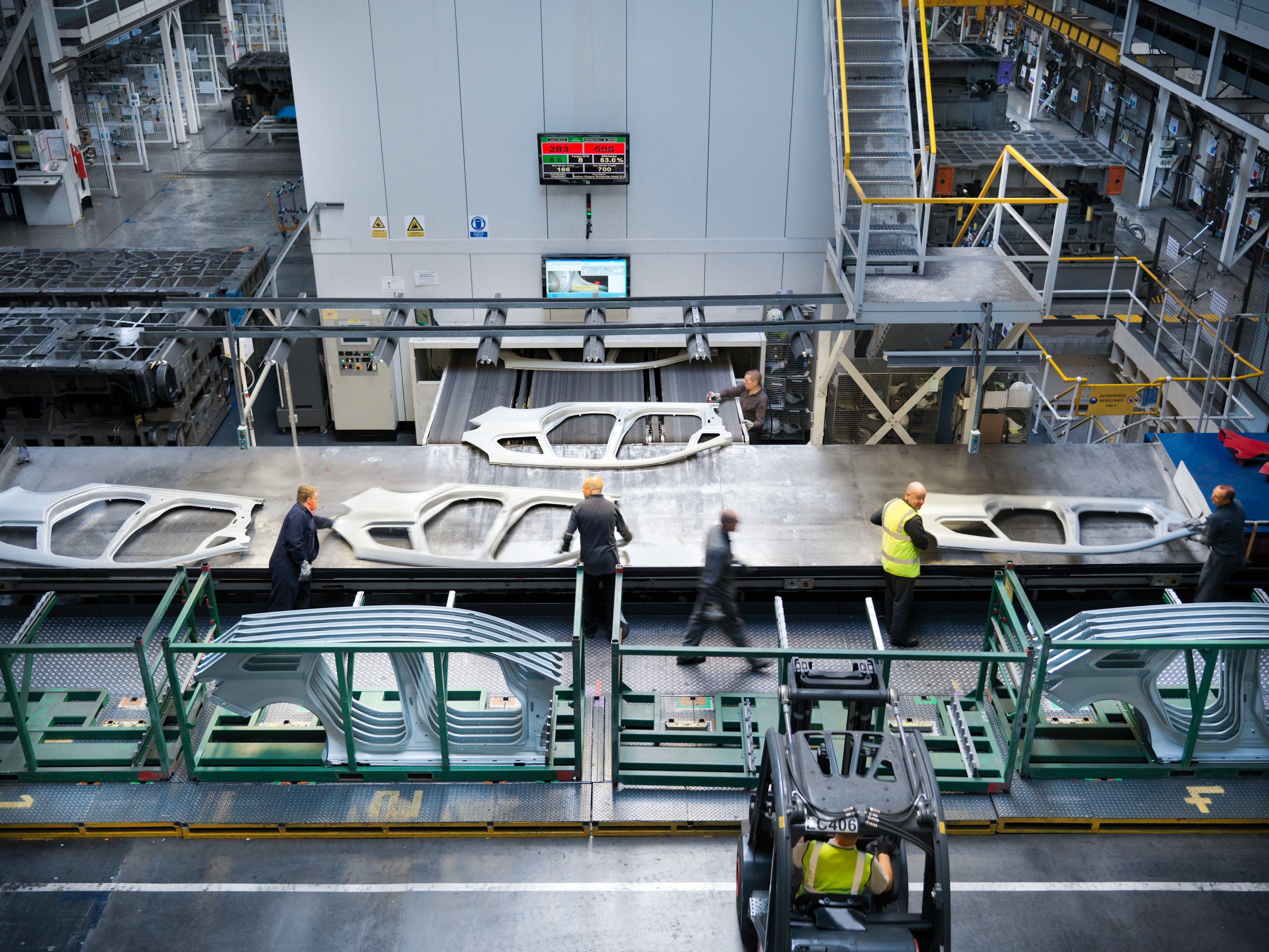 Workers in a factory inspect large metal car parts on a conveyor system.
