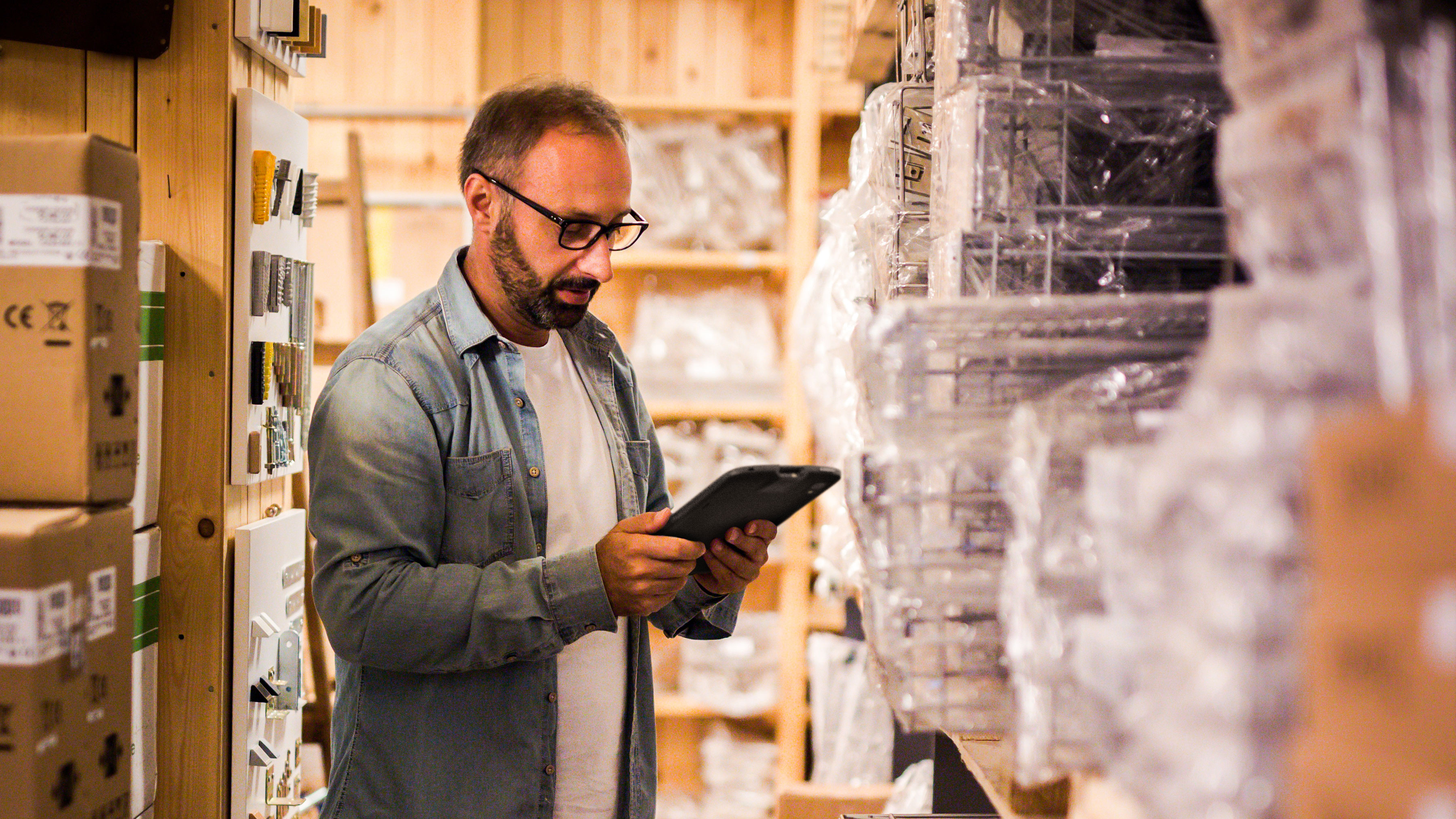A back-of-house employee is standing in a narrow aisle of a store room, holding a tablet and looking at shelves filled with various plastic-wrapped items.