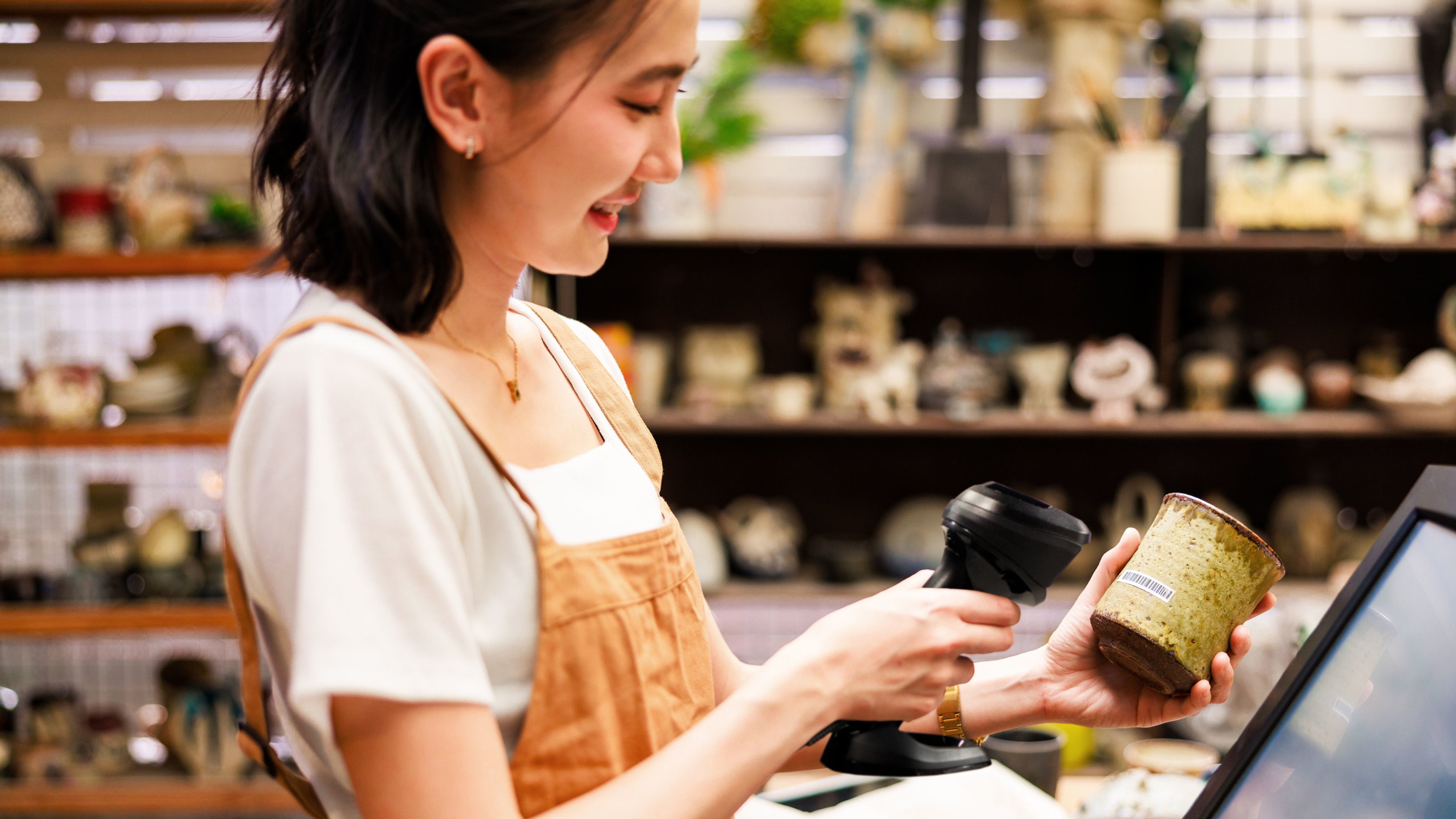 A front-of-house employee using a handheld barcode scanner to ring up a ceramic planter during checkout inside a garden center.