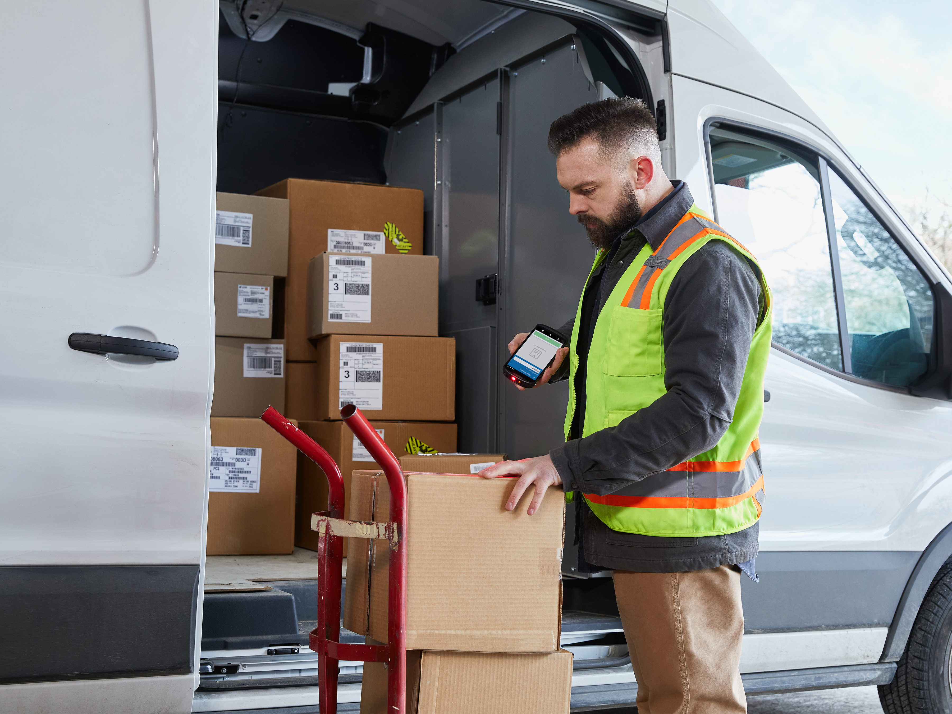 Delivery person next to an open van using a mobile computer to scan a shipping label on a box. 
