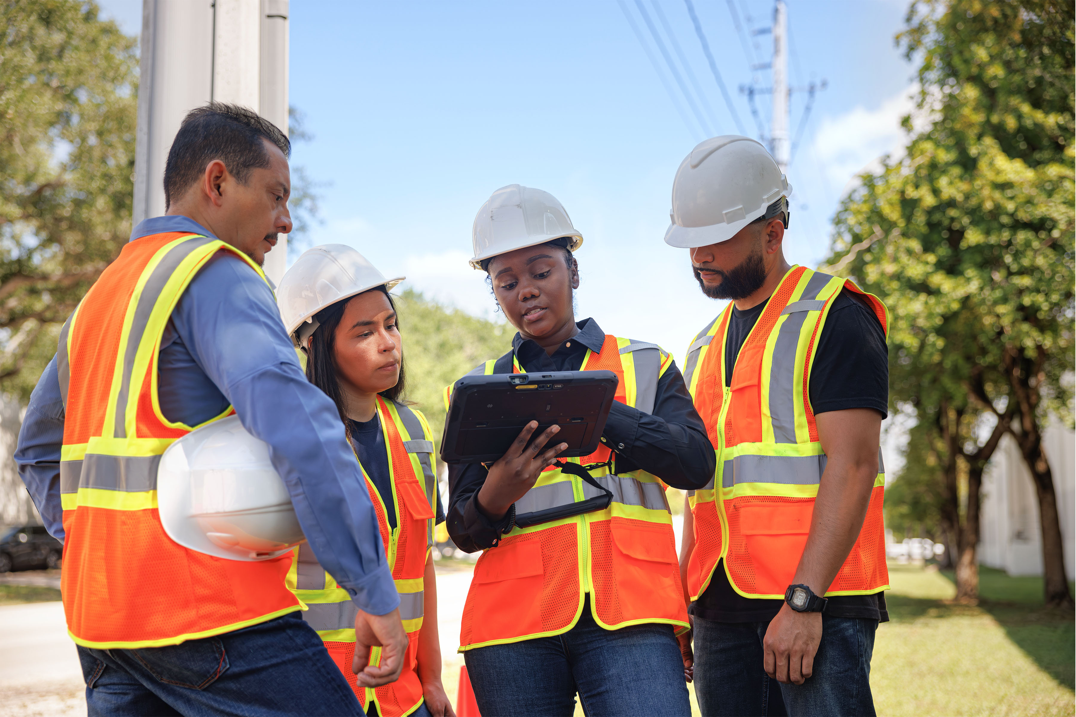 Utility workers with orange safety vests and white hard hatsoutside reviewing a tablet screen together