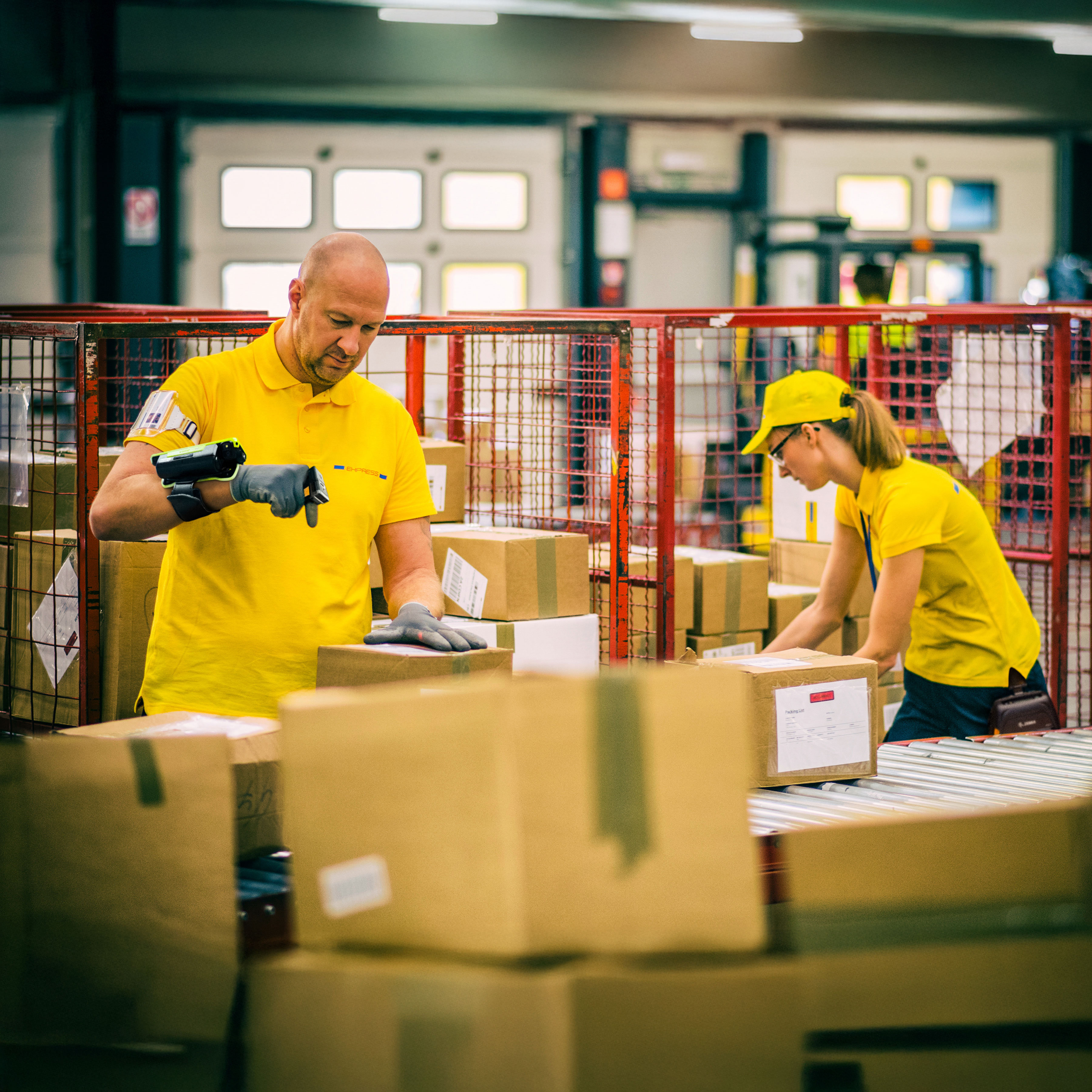 Two postal workers in yellow uniforms are sorting and scanning boxes using wearable scanners.