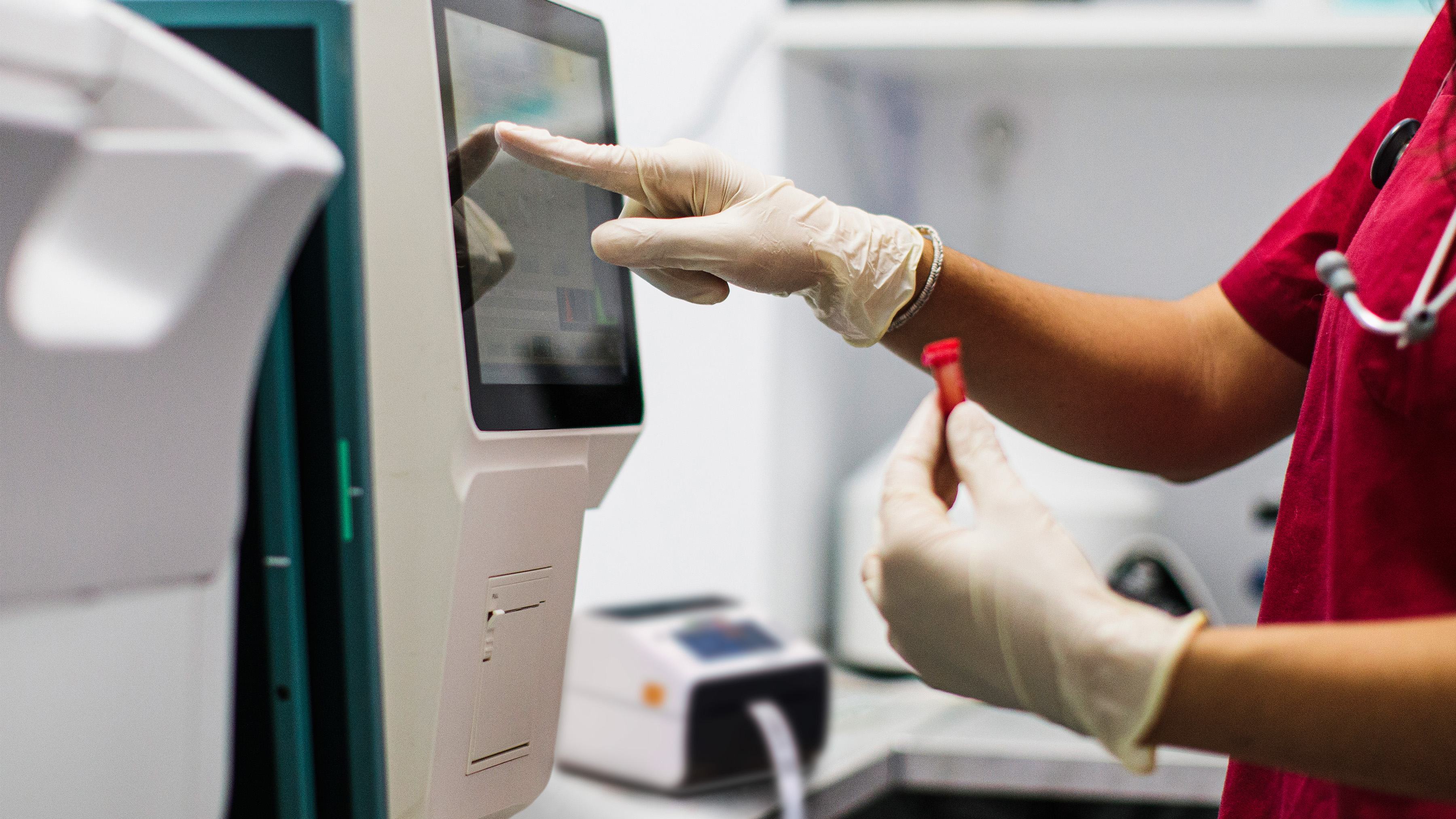 A healthcare worker in red with gloved hands in a healthcare setting using a touchscreen while holding a blood sample vial with a desktop printer in the background.