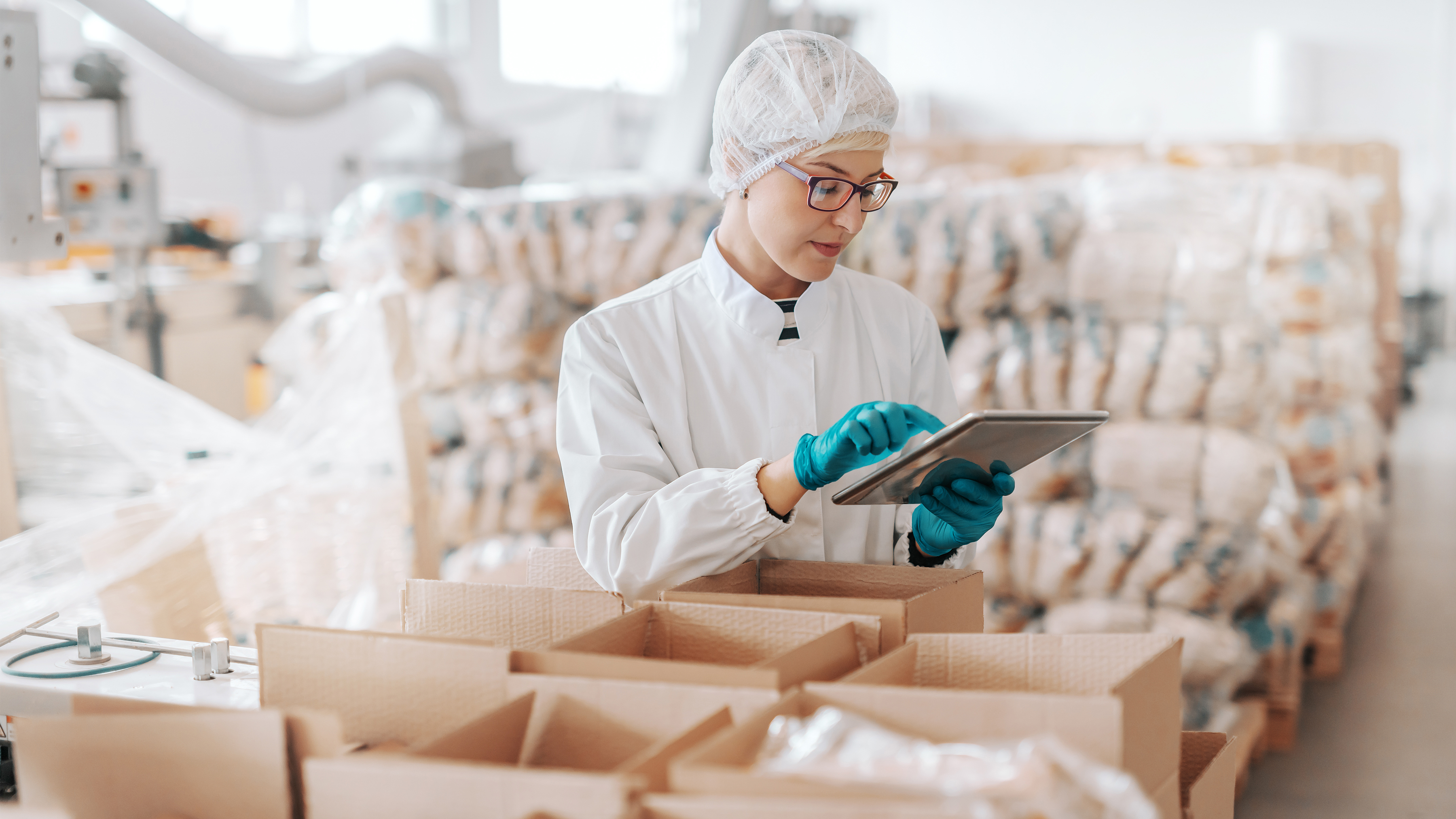 A woman in scrubs and blue gloves working on a mobile tablet, surrounded by open boxes and packages.