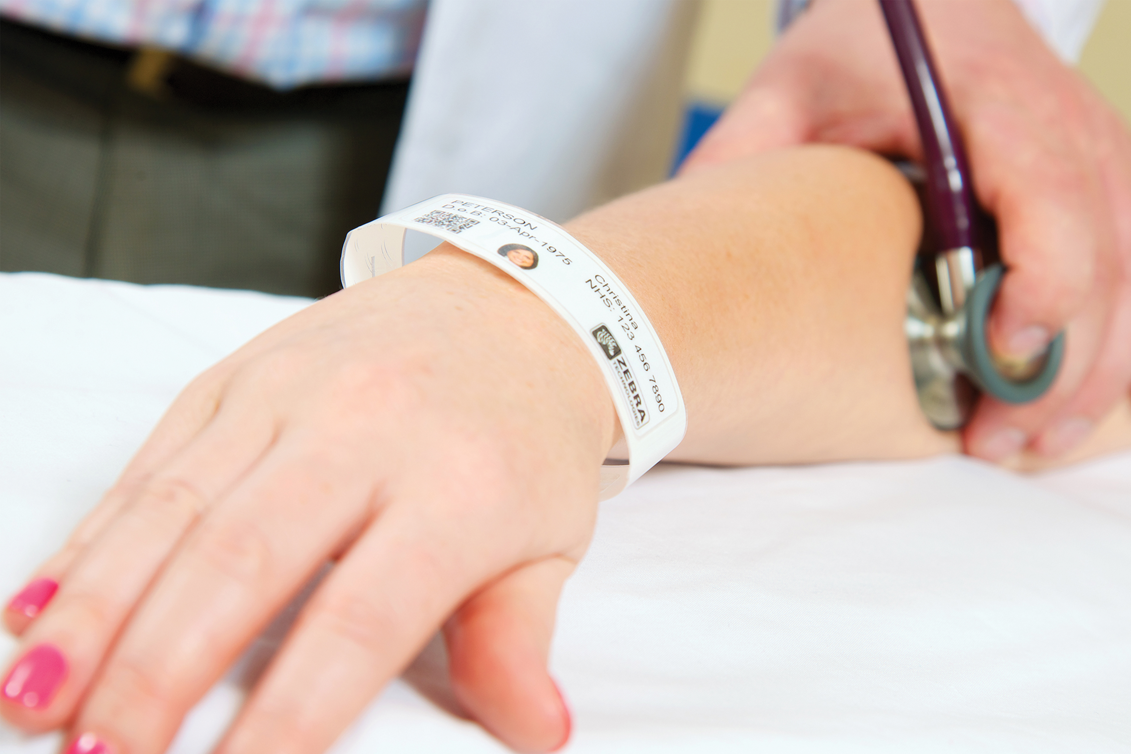 A patient's arm wearing a printed wristband, while a doctor is using stethoscope to check vitals.