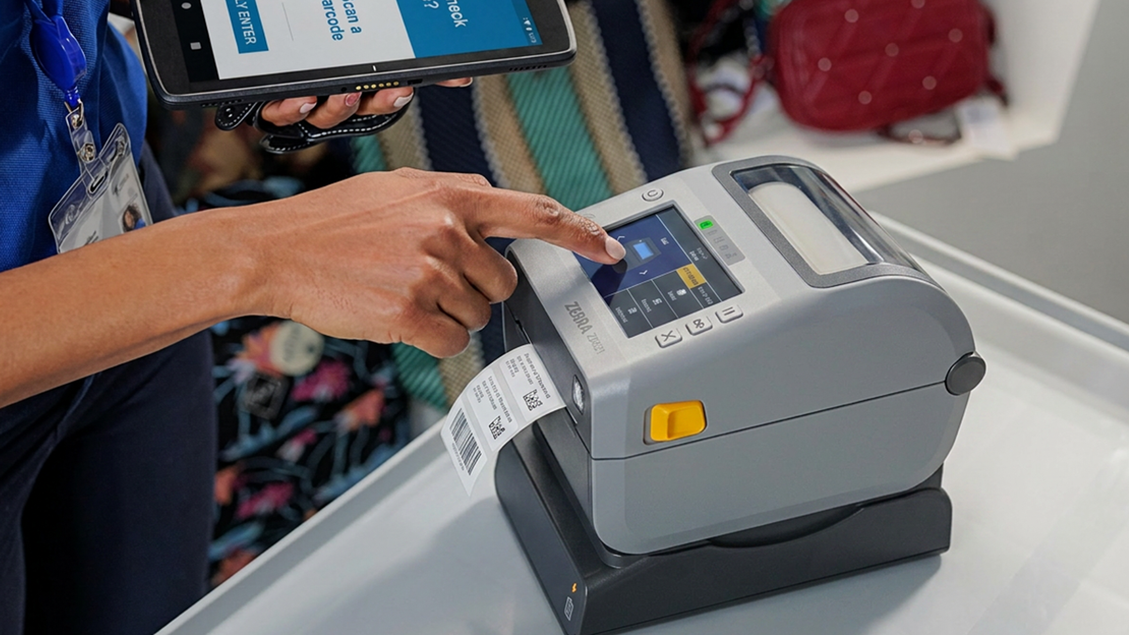 A close-up image showing a retail or logistics associate using a Zebra ZD621 desktop label printer. The user is interacting with the printer's color touchscreen with one hand while holding a Zebra mobile computer in the other, highlighting an efficient, modern workflow. A fresh barcode label is being printed.
