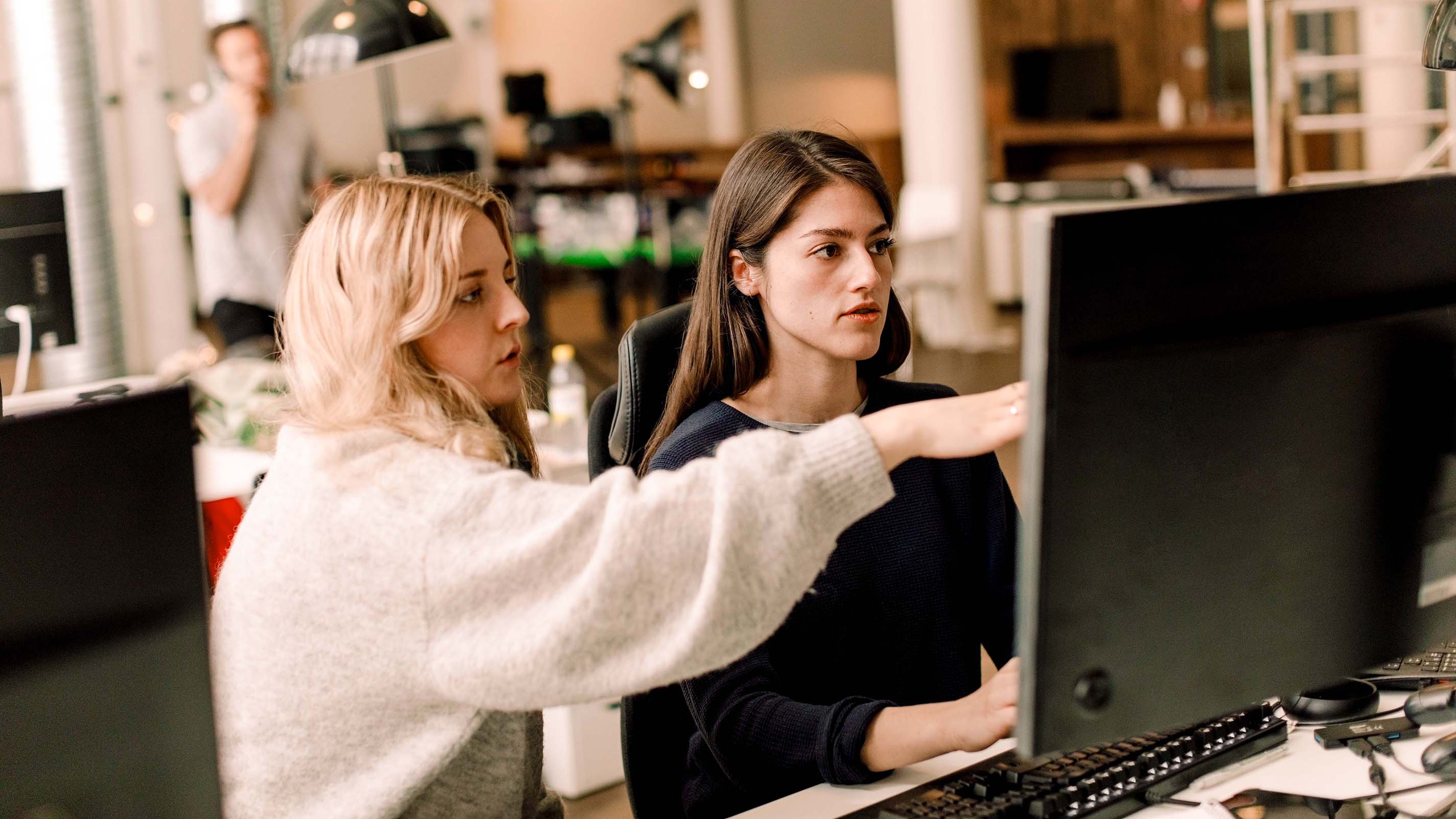 Two colleagues are working together at a desktop computer in an office, with one person pointing at the screen and explaining a topic.