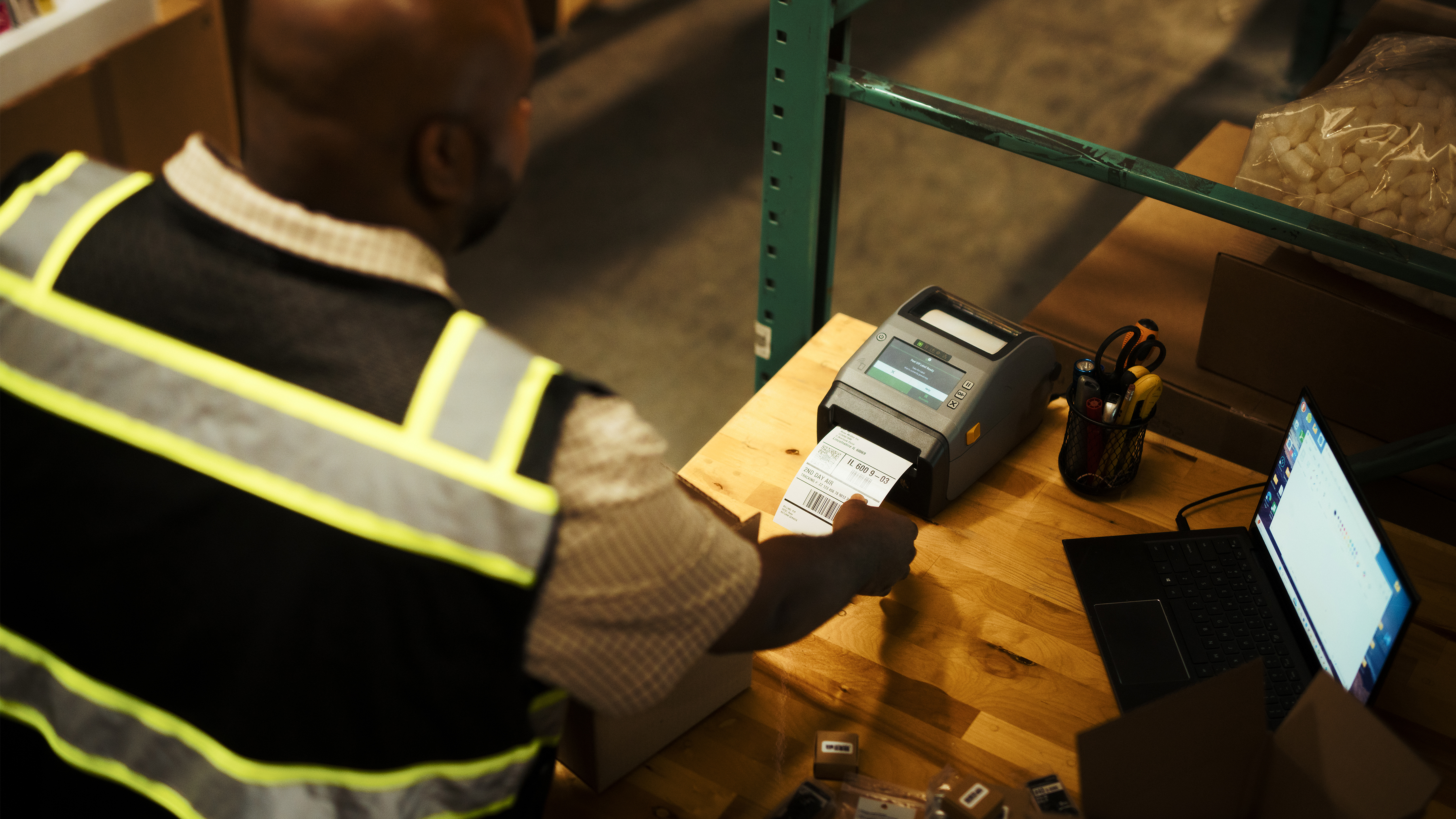 Overhead view of a man in a warehouse with safety vest at a workstation with a laptop open, printing a packing label with a desktop printer.