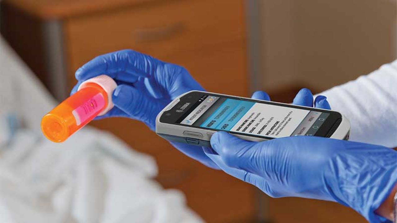 A clinician wearing gloves uses a Zebra TC-52HC mobile computer to scan a prescription bottle label