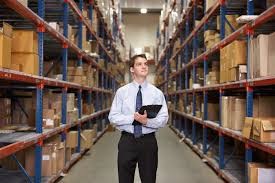 A man stands in the middle of a warehouse aisle with a tablet in hand, looking up at the inventory on the hshelves.